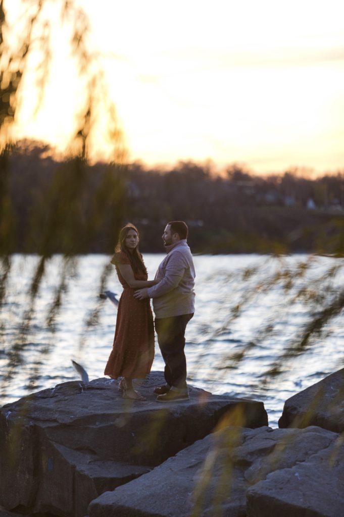 couple standing on rocks by a lake at sunset with tree branches in the foreground