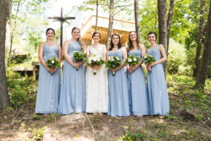 Bridesmaids holding white rose bouquets in a forest with a bride