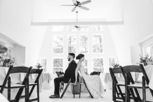 A bride and groom sit in chairs in a foyer and hold hands while praying back to back