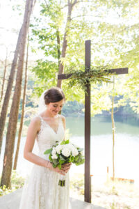 Bride in a forest by a lake looking at her roses with a cross in the background