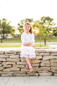 A flower girl wearing a white robe is sitting on a stone wall during sunrise in front of trees