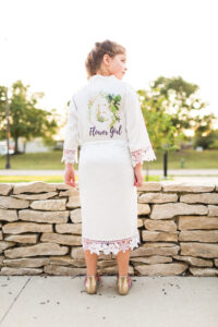 A flower girl is standing in front of a stone wall during sunrise with trees in the distance