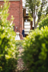 Couple holding hands around the corner of a brick building between two bushes