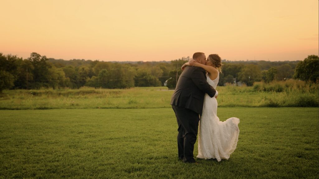bride and groom embracing and kissing at sunset with leg popped up, trees and tall grass in background at upscale barn wedding venue stone valley meadows
