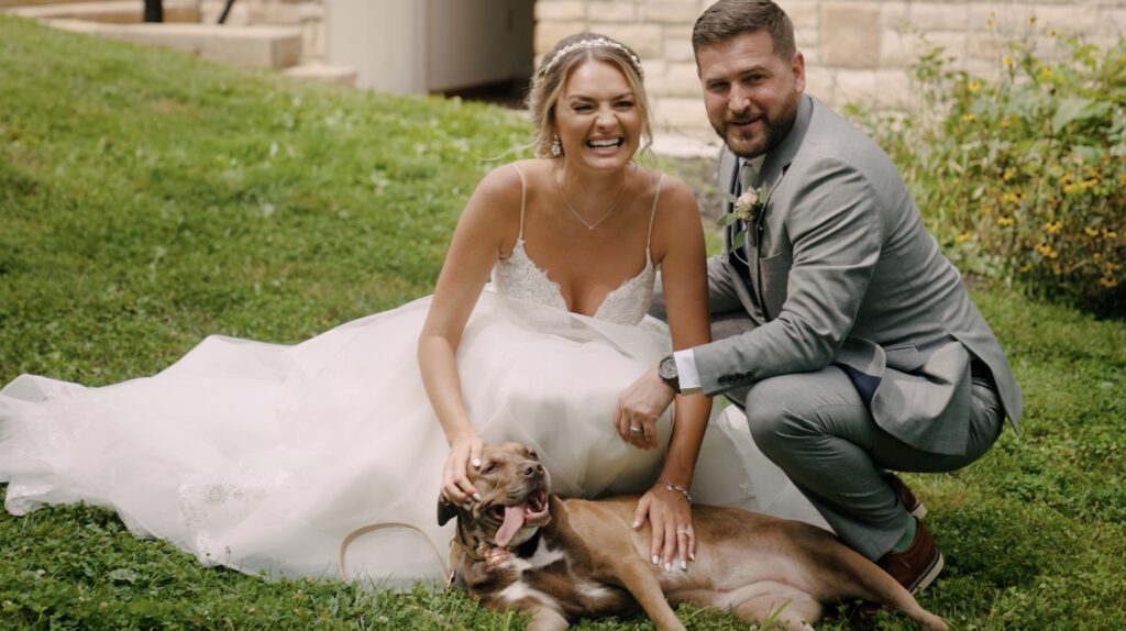 Bride and groom patting and petting dog in wedding dress and suit while posing for a family photo and smiling at Ohio Whitetail Lodge