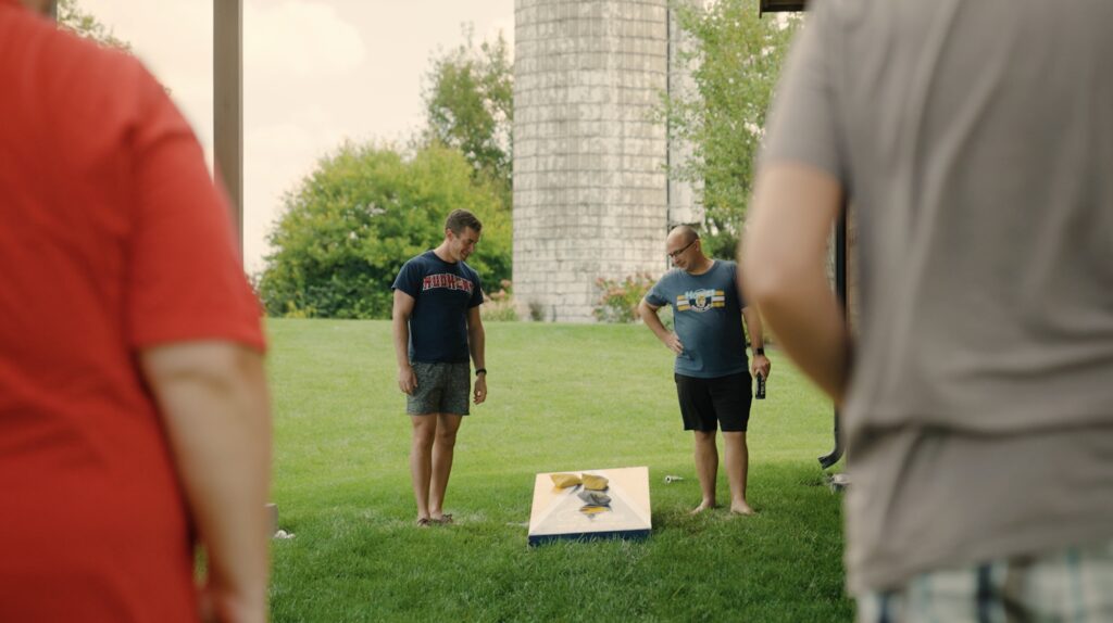 Guys staring down at a cornhole board outside as a beanbag lands on the board