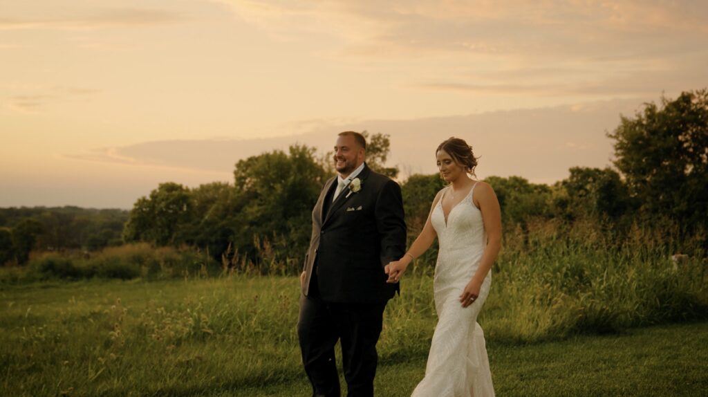 bride and groom walking while holding hands at sunset with tall grasses and fields and trees behind them at stone valley meadows for their upscale barn wedding