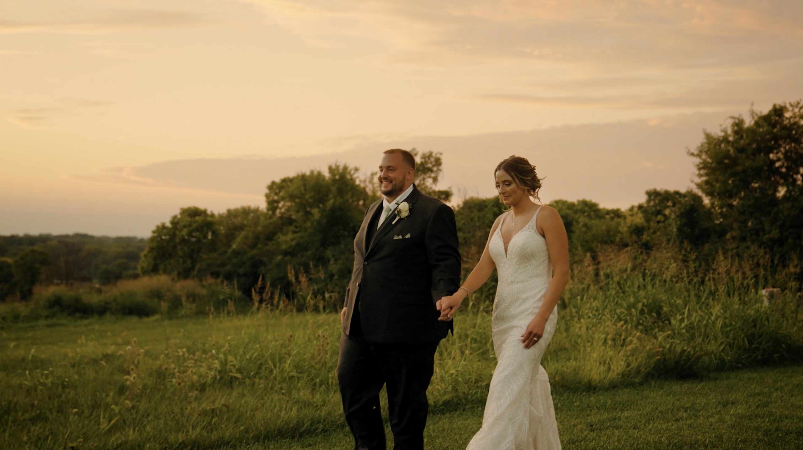 Bride and groom smiling while walking together holding hands at sunset in front of trees and tall grass
